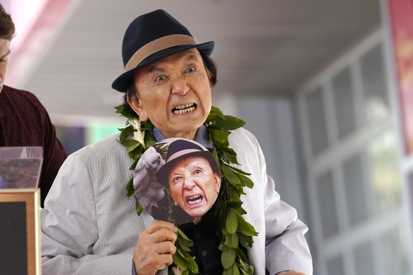 An actor holds a paper fan with his own snarling face on it and snarls jokingly.